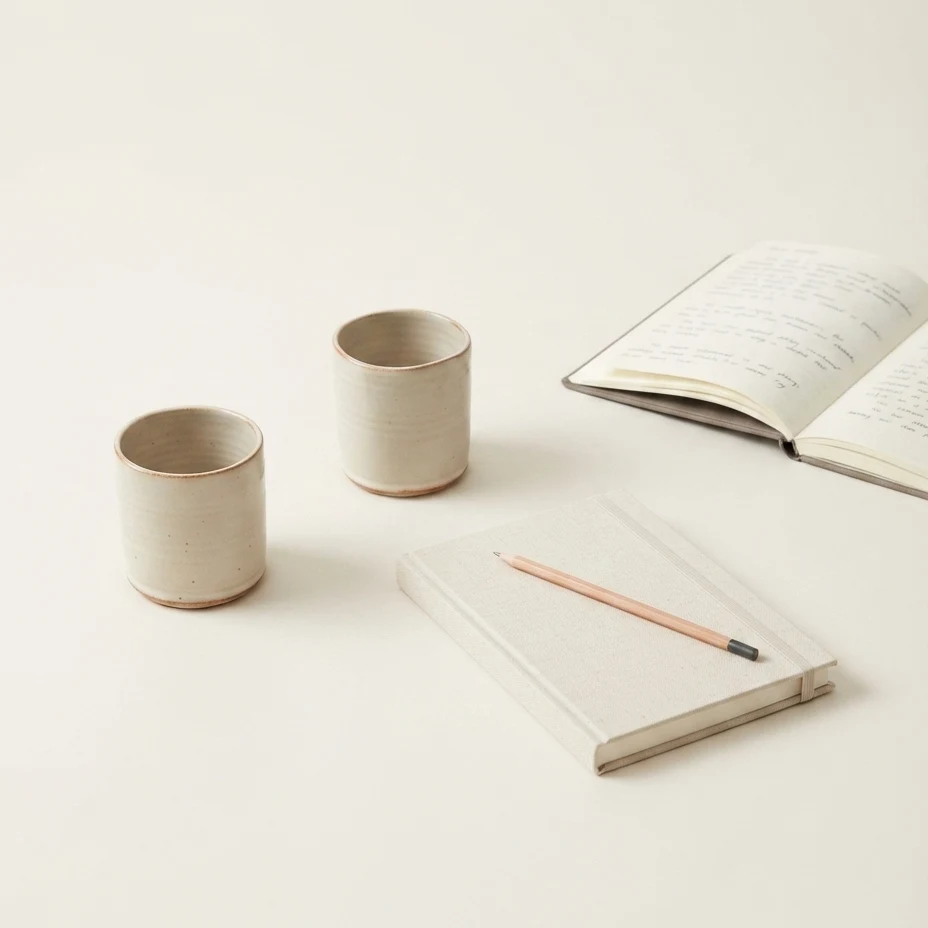 Two stoneware cups and a closed notebook with a plain pencil resting on it. Behind them, an open notebook angles away, its handwriting soft and unreadable.